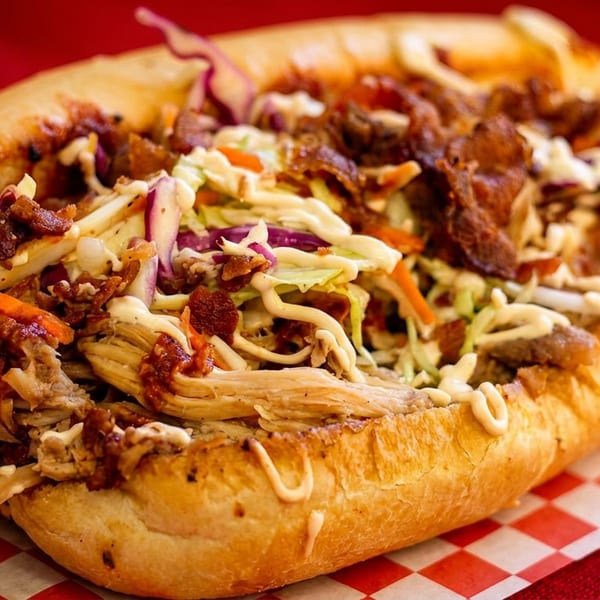 A large pulled pork sandwich with coleslaw, bacon bits, and sauce on a red and white checkered paper.
