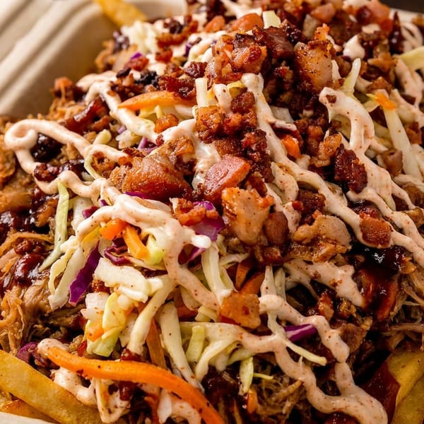 Close-up of loaded fries with pulled pork, coleslaw, bacon bits, and a creamy sauce in a takeout container.