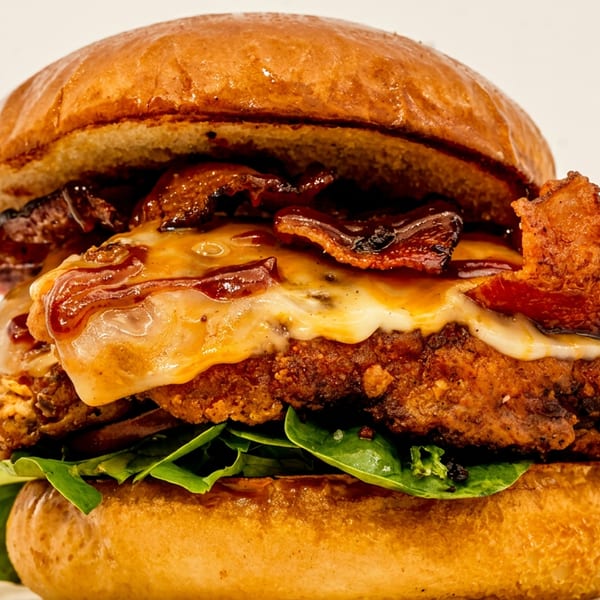 Close-up of a fried chicken sandwich with melted cheese, bacon, and greens on a brioche bun.