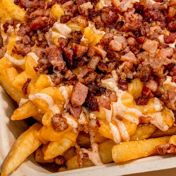 Close-up of golden french fries topped with melted cheese, bacon bits, and a creamy sauce in a paper tray.