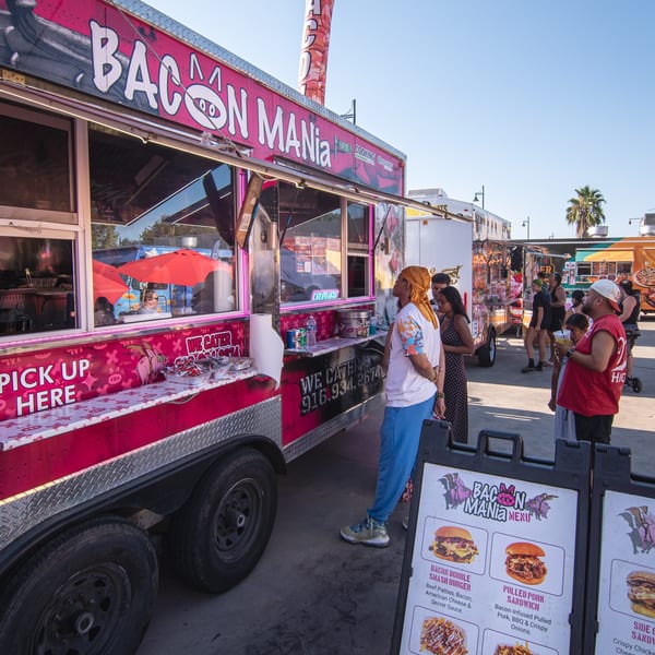 A pink "Bacon Mania" food truck with a wavy inflatable tube man and people ordering food.